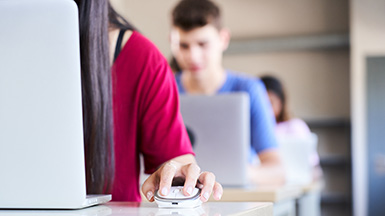 Close-up of a hand clutching a mouse in a high school classroom, a group of cheerful students study with a laptop computer. Concept of technology and education